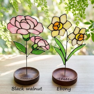May include: Two stained glass flower sculptures. One features pink carnations, the other yellow daffodils. Each flower is mounted on a dark wooden base. The carnation base is labeled "Vivian", and the daffodil base is labeled "Stella". The base materials are labeled "Black walnut" and "Ebony".
