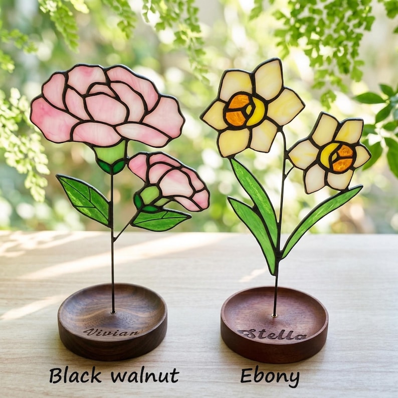 May include: Two stained glass flower sculptures. One features pink carnations, the other yellow daffodils. Each flower is mounted on a dark wooden base. The carnation base is labeled "Vivian", and the daffodil base is labeled "Stella". The base materials are labeled "Black walnut" and "Ebony".