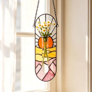 Puede incluir: Un colgante de pared de vitral vertical que presenta un paisaje de atardecer con paneles naranjas, amarillos y rosas. Un tubo de ensayo de vidrio transparente está montado en el centro, sosteniendo un pequeño ramo de margaritas blancas con centros amarillos.