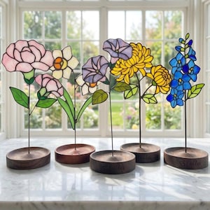 May include: A selection of stained glass flowers in shades of pink, yellow, blue, and purple. Each flower is mounted on a dark wooden base. The bases are engraved with the name "Stella".