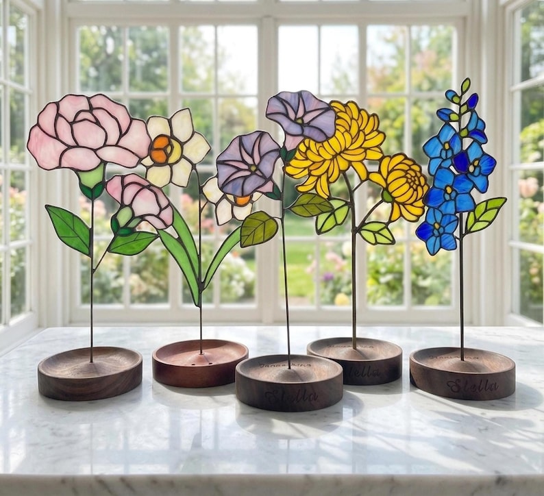 May include: A selection of stained glass flowers in shades of pink, yellow, blue, and purple. Each flower is mounted on a dark wooden base. The bases are engraved with the name "Stella".