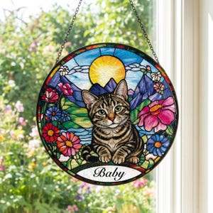 Puede incluir: Vidriera redonda con un gato atigrado y la palabra "Baby" debajo. La obra de arte incluye un paisaje montañoso, flores y un sol. Diseñado para colgar con una cadena.