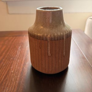 May include: A cylindrical ceramic vase with a textured, ribbed exterior in a warm terracotta brown. The upper portion features a smooth, light beige glaze that drips down over the ribbed body, creating a distinct contrast in both color and texture.
