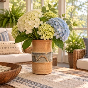May include: A cylindrical ceramic vase with a tan glazed finish and a central dark gray band featuring embossed wave-like patterns. The vase is filled with a large bouquet of white, light green, and pale blue hydrangea flower clusters.
