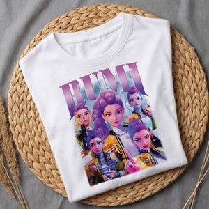 May include: White t-shirt with a graphic of five stylized women with purple hair and yellow jackets. The word "RUMI" is printed in large, purple letters across the top of the design. The shirt is displayed on a woven, circular mat.