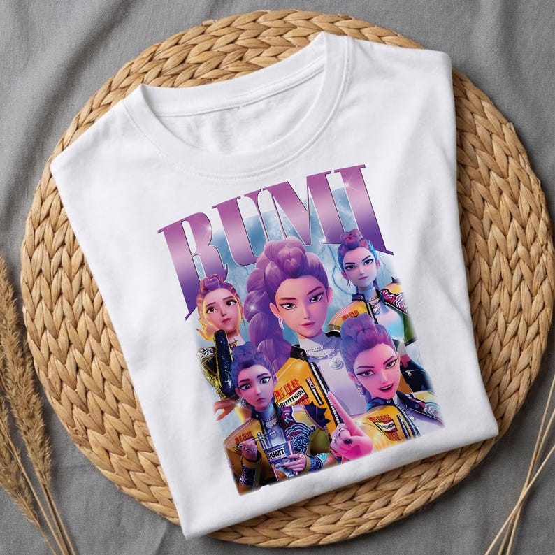 May include: White t-shirt with a graphic of five stylized women with purple hair and yellow jackets. The word "RUMI" is printed in large, purple letters across the top of the design. The shirt is displayed on a woven, circular mat.