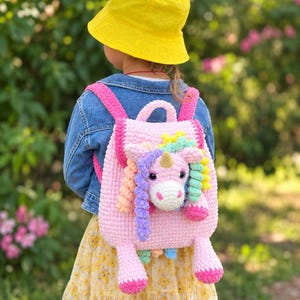 May include: A light pink crocheted backpack featuring a three-dimensional unicorn head on the front. The unicorn has a gold horn, dark eyes, and a multi-colored mane made of yarn curls. The backpack has bright pink straps and two dangling legs.