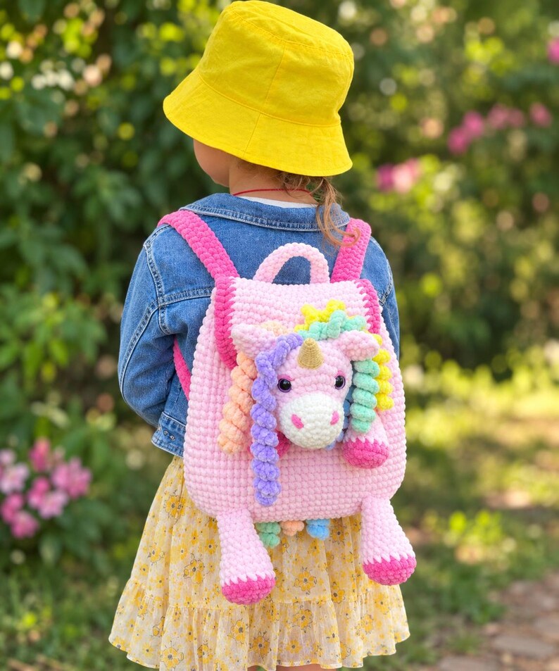 May include: A light pink crocheted backpack featuring a three-dimensional unicorn head on the front. The unicorn has a gold horn, dark eyes, and a multi-colored mane made of yarn curls. The backpack has bright pink straps and two dangling legs.