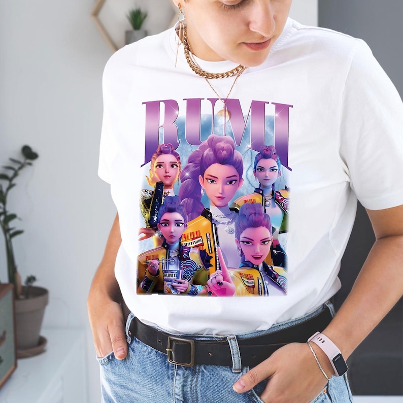 May include: White t-shirt with a graphic of the word "RUMI" in purple, and a group of animated female characters. The characters are wearing yellow and black jackets. The t-shirt is made of a soft, comfortable material.