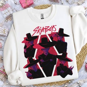 May include: White crewneck sweatshirt featuring a graphic of five figures in black outfits and wide-brimmed hats. The figures are set against a red background with a geometric design and the text "SAJA BOYS" in red.