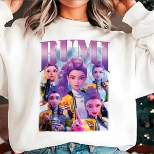 May include: White sweatshirt with a graphic of several animated women with purple hair and yellow jackets. The word "RUMI" is printed in large, purple letters above the image. The design has a retro, cartoon style.