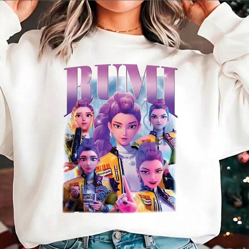 May include: White sweatshirt with a graphic of several animated women with purple hair and yellow jackets. The word "RUMI" is printed in large, purple letters above the image. The design has a retro, cartoon style.