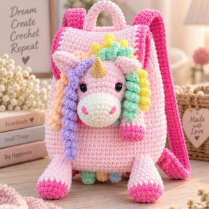 May include: A pink crochet backpack featuring a three-dimensional unicorn head on the front. The unicorn has a gold horn, black eyes, and a multi-colored curly mane in shades of yellow, green, and purple. The bag has two pink shoulder straps.