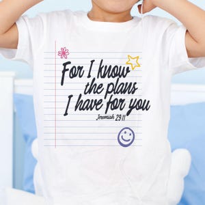 K&ouml;nnte beinhalten: Ein wei&szlig;es Kurzarm-T-Shirt mit einer Grafik von liniertem Notizbuchpapier und dem schwarzen Text "For I know the plans I have for you" sowie "Jeremiah 29:11". Das Design enth&auml;lt eine rosa Blume, einen gelben Stern und ein lila Smiley-Gesicht.