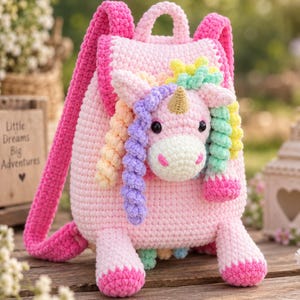 May include: A light pink crochet backpack featuring a three-dimensional unicorn head on the front flap. The unicorn has a gold horn, black eyes, and a multi-colored mane in pastel shades of purple, orange, yellow, and green. It has two pink straps.