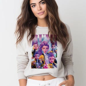 May include: A light grey crew neck sweatshirt with a graphic of a group of animated women, with the word "RUMI" in purple. The women wear yellow jackets and have different hairstyles. The sweatshirt is a casual style.