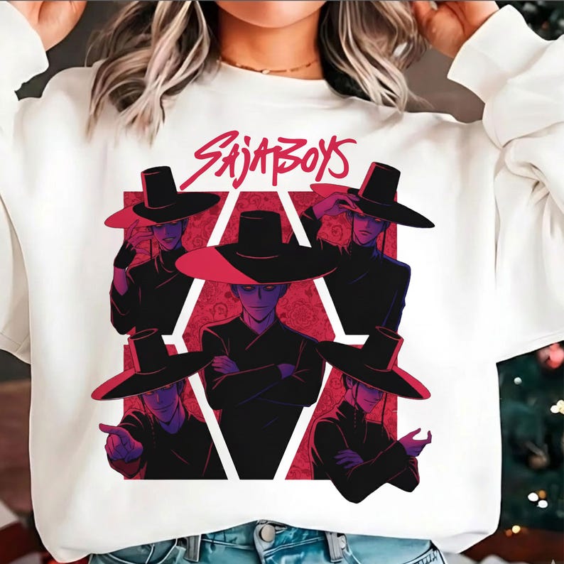 May include: White sweatshirt featuring a graphic of stylized figures wearing wide-brimmed hats. The figures are set against a red geometric background with the text "SAJA BOYS" in red above the figures.