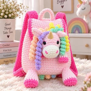 May include: A pink crocheted backpack featuring a three-dimensional unicorn head on the front flap. The unicorn has a gold horn, black eyes, and a multi-colored mane in shades of purple, yellow, and green. The backpack has two shoulder straps.