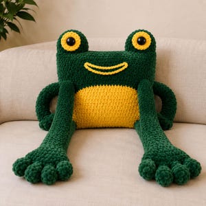 May include: A dark green and yellow crocheted frog plush toy with a rectangular body, long arms, and large eyes. The frog features a bright yellow belly, a yellow stitched smile, and two prominent black eyes with yellow borders on top of its head.