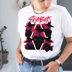May include: White t-shirt featuring a graphic design with the text "SAJA BOYS" in red, above a stylized illustration of figures wearing black hats and outfits, set against a red geometric background.