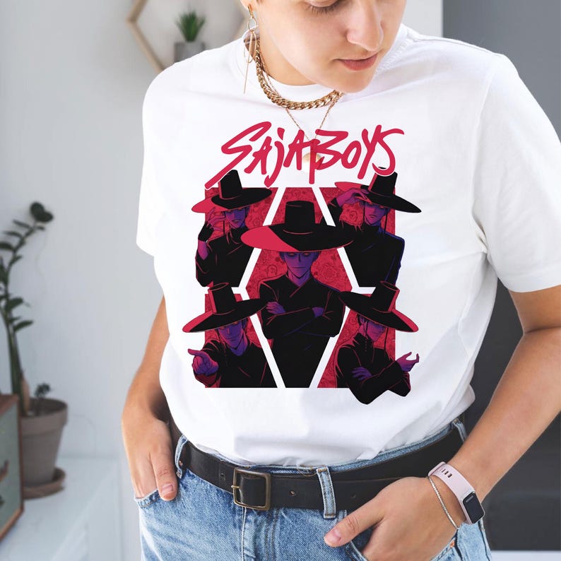 May include: White t-shirt featuring a graphic design with the text "SAJA BOYS" in red, above a stylized illustration of figures wearing black hats and outfits, set against a red geometric background.