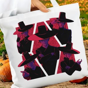 May include: White tote bag featuring a graphic of five figures in black outfits and wide-brimmed hats. The figures are set against a red patterned background within a white geometric shape. The artwork has a manga or anime style.
