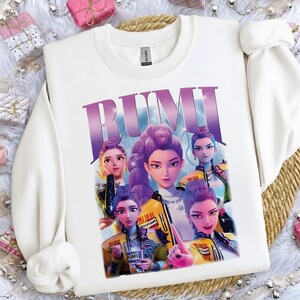 May include: White sweatshirt with a graphic of five animated women with purple hair and yellow jackets. The word "RUMI" is printed in large, purple letters across the top of the design. The sweatshirt has a crew neck and long sleeves.