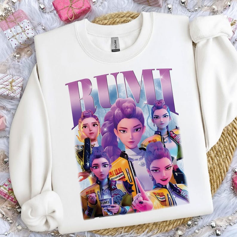 May include: White sweatshirt with a graphic of five animated women with purple hair and yellow jackets. The word "RUMI" is printed in large, purple letters across the top of the design. The sweatshirt has a crew neck and long sleeves.