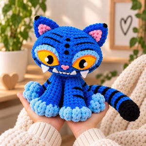 Può includere: Un peluche a forma di gatto lavorato all'uncinetto in blu con strisce nere, grandi occhi gialli, un piccolo naso rosa e prominenti zanne bianche. Il gatto presenta accenti azzurri sulle orecchie e sulle zampe e una lunga coda a strisce.