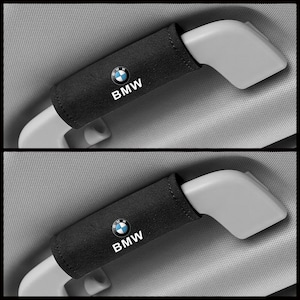 Könnte beinhalten: Zwei schwarze Autosicherheitsgurtbezüge mit dem BMW-Logo in Weiß und Blau. Die Bezüge sind um die Sicherheitsgurtschnalle gewickelt. Der Hintergrund ist ein hellgrauer Autoinnenraum.