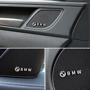 May include: Close-up of a car door panel featuring a speaker grill with the BMW logo. The speaker grill is black with a mesh pattern, and the BMW logo is white. The door panel is dark gray.