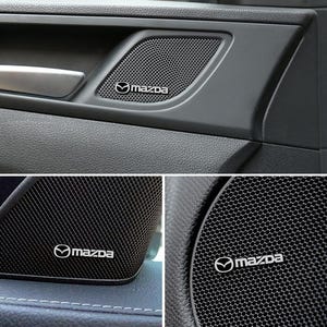 Może przedstawiać: Czarny panel drzwi samochodu z kratką głośnika i białym logo Mazda. Głośnik jest osadzony we wnęce panelu drzwi. Widoczna jest również srebrna klamka.