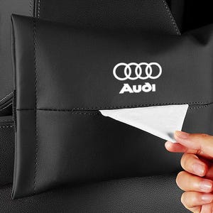 Puede incluir: Soporte de pañuelos de cuero negro con el logotipo de Audi en blanco. El soporte está sujeto al reposacabezas del asiento del coche. Una mano saca un pañuelo blanco del dispensador.