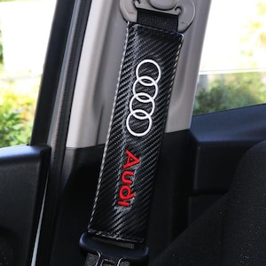 Può includere: Una copertura nera per cintura di sicurezza con un motivo in fibra di carbonio. La copertura presenta il logo Audi in bianco e la scritta "Audi" in rosso. La copertura è attaccata a una cintura di sicurezza nera.