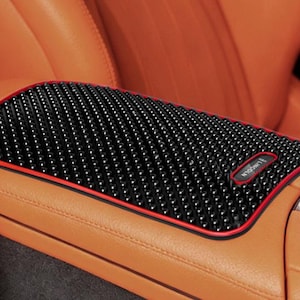 May include: Black textured car console cover with a red trim, placed on a tan leather car seat. The cover has a grid-like pattern and a small red and white logo. The car interior is tan and black.