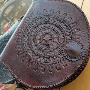 May include: A brown leather saddlebag with a circular, embossed design. The bag features a detailed, symmetrical pattern with a central starburst and a smaller, decorative circle on the side. A dark brown strap is attached.
