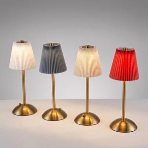 Puede incluir: Cuatro lámparas de mesa con bases circulares y tallos delgados en tono latón. Cada lámpara presenta una pantalla cónica plisada en blanco, gris oscuro, beige claro y rojo. Las lámparas están alineadas, con la primera y la tercera encendidas.