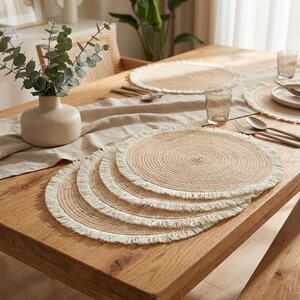 Peut inclure: Un ensemble de sets de table ronds tressés avec un centre beige naturel et une bordure à franges blanches. Les sets sont empilés sur une table en bois, accompagnés d'un chemin de table beige, d'un vase en céramique avec de l'eucalyptus et de verres.