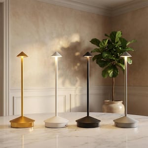 Peut inclure: Quatre lampes de table sans fil fines alignées sur une surface en marbre. Chaque lampe présente une base conique, une tige verticale fine et un petit abat-jour pointu émettant une lueur douce et chaude. Finitions or, blanc, noir et gris.