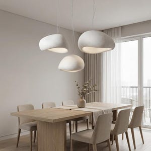 Peut inclure: Trois suspensions blanches en forme de galet organique sont suspendues par de fins cordons au-dessus d'une table à manger rectangulaire en bois. Les lampes présentent une finition mate lisse avec une ouverture inférieure diffusant une lumière douce.