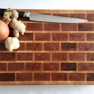 May include: A rectangular end-grain chopping board with a brick-like pattern, featuring alternating dark and light wood blocks. A large chef's knife, an onion, a bulb of garlic, and ginger are on the board.