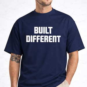 Könnte beinhalten: Marineblaues T-Shirt mit dem weißen Schriftzug "BUILT DIFFERENT". Das kurzärmelige Oberteil hat einen Rundhalsausschnitt und ist aus einem weichen Stoff gefertigt. Ein lässiges Kleidungsstück für den Alltag.