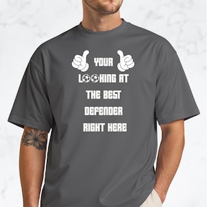 Puede incluir: Camiseta gris con texto blanco y gráficos de pulgares arriba. El texto dice "YOUR LOOKING AT THE BEST DEFENDER RIGHT HERE" con gráficos de balones de fútbol. La camiseta es de estilo casual.