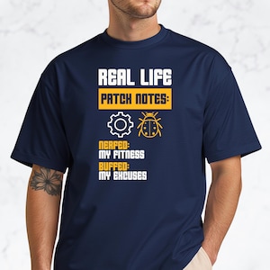 May include: Navy blue t-shirt featuring white and yellow text. The text says "REAL LIFE PATCH NOTES: NERFED: MY FITNESS, BUFFED: MY EXCUSES" with gear and ladybug graphics. A casual tee for everyday wear.