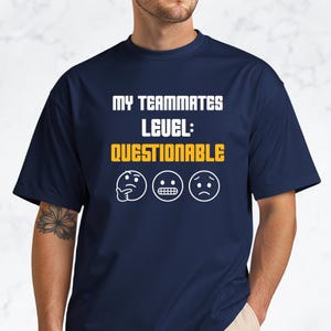 May include: A navy blue t-shirt featuring the text "MY TEAMMATES LEVEL: QUESTIONABLE" in white and yellow. The design includes three emoji faces. The t-shirt has a crew neck and short sleeves.