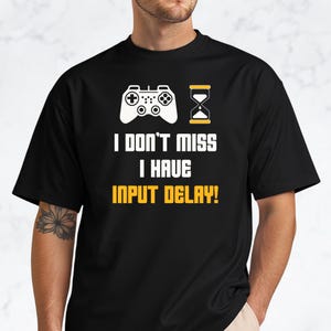 May include: Black t-shirt with a white graphic of a game controller and an hourglass. The text on the shirt reads "I DON'T MISS I HAVE INPUT DELAY!" in white and yellow block letters.