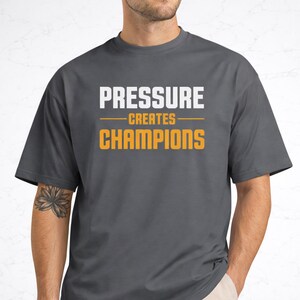 Könnte beinhalten: Ein dunkelgraues T-Shirt mit dem Text "PRESSURE CREATES CHAMPIONS" in Weiß und Orange. Der Text ist mittig auf der Vorderseite des Shirts platziert.