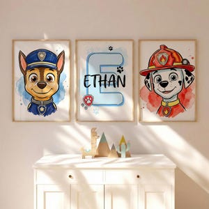 Puede incluir: Tres impresiones en acuarela enmarcadas. Una muestra un perro policía con uniforme azul, otra muestra el nombre "Ethan" con una huella de pata, y la tercera representa un dálmata con uniforme de bombero. Las impresiones están enmarcadas.