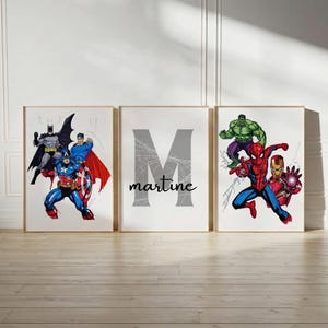 May include: A set of three wall art prints featuring comic book characters. The left print shows Batman, Superman, and Captain America. The centre print displays a large grey letter M with the name martine written below. The right print features Hulk, Spider-Man, and Iron Man.