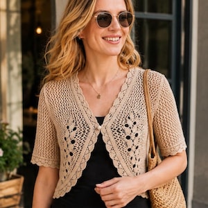 May include: A beige crochet cardigan featuring an open-knit pattern with leaf-like motifs, short sleeves, and a scalloped edge. The garment is secured at the center with a single white button and is worn over a plain black top.